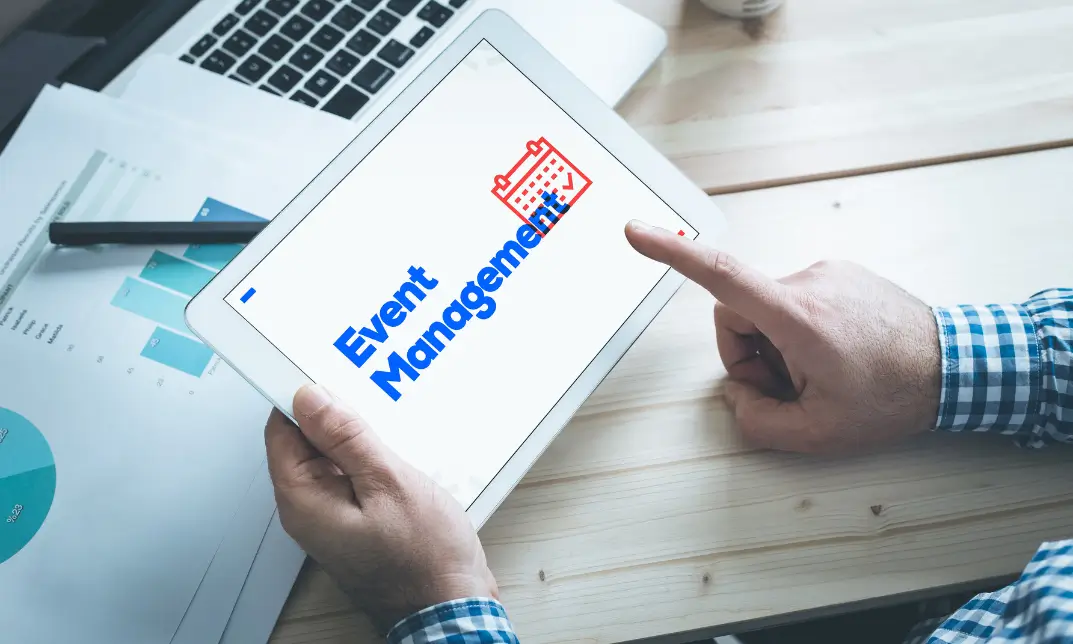 Tablet screen with Event Management text and calendar icon, held over a desk with papers and a pen.
